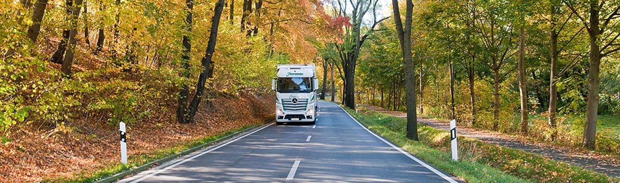 Weißer LKW fährt auf einer von Herbstbäumen gesäumten Straße.