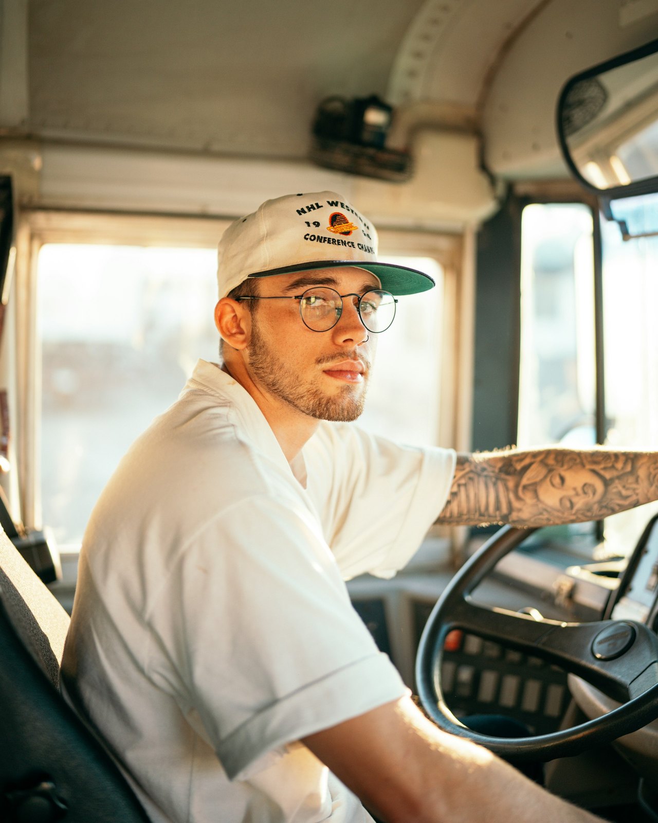Ein junger Mann mit Brille und Baseballkappe sitzt am Steuer eines Busses und blickt zur Seite.