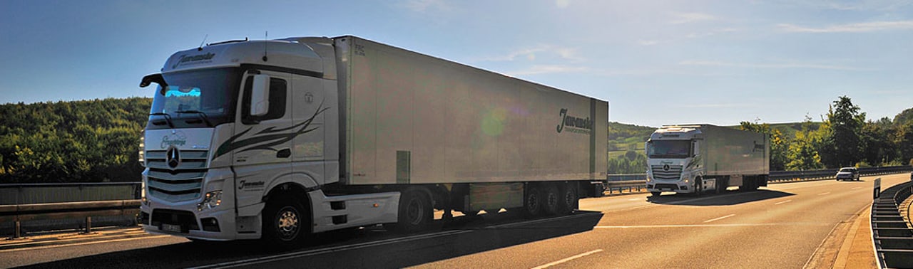 Zwei weiße Lastwagen fahren auf einer sonnigen Autobahn, umgeben von grünen Bäumen.
